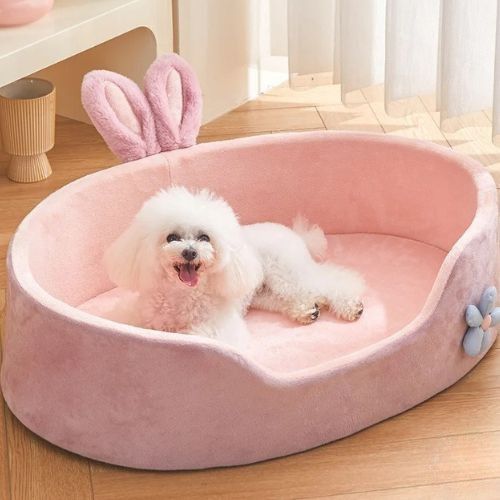 Petit chien blanc allongé dans un panier pour chien rose avec oreilles décoratives, installé dans un salon lumineux au sol en bois.
