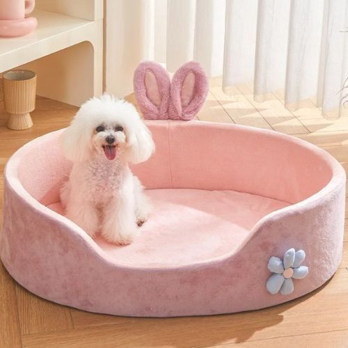 Petit chien blanc assis dans un panier pour chien rose en tissu doux, avec oreilles décoratives et fleur cousue, placé dans un salon lumineux.
